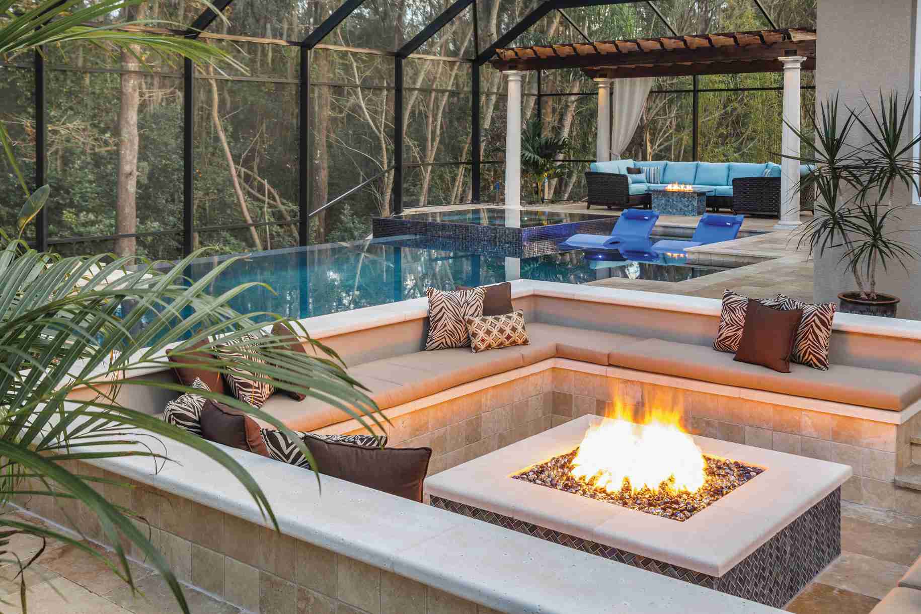 Luxurious outdoor living space featuring a swimming pool with submerged blue lounge chairs, a built-in fire pit surrounded by a U-shaped seating area with beige cushions, and a separate lounge under a pergola, all within a screened enclosure overlooking a dense forest